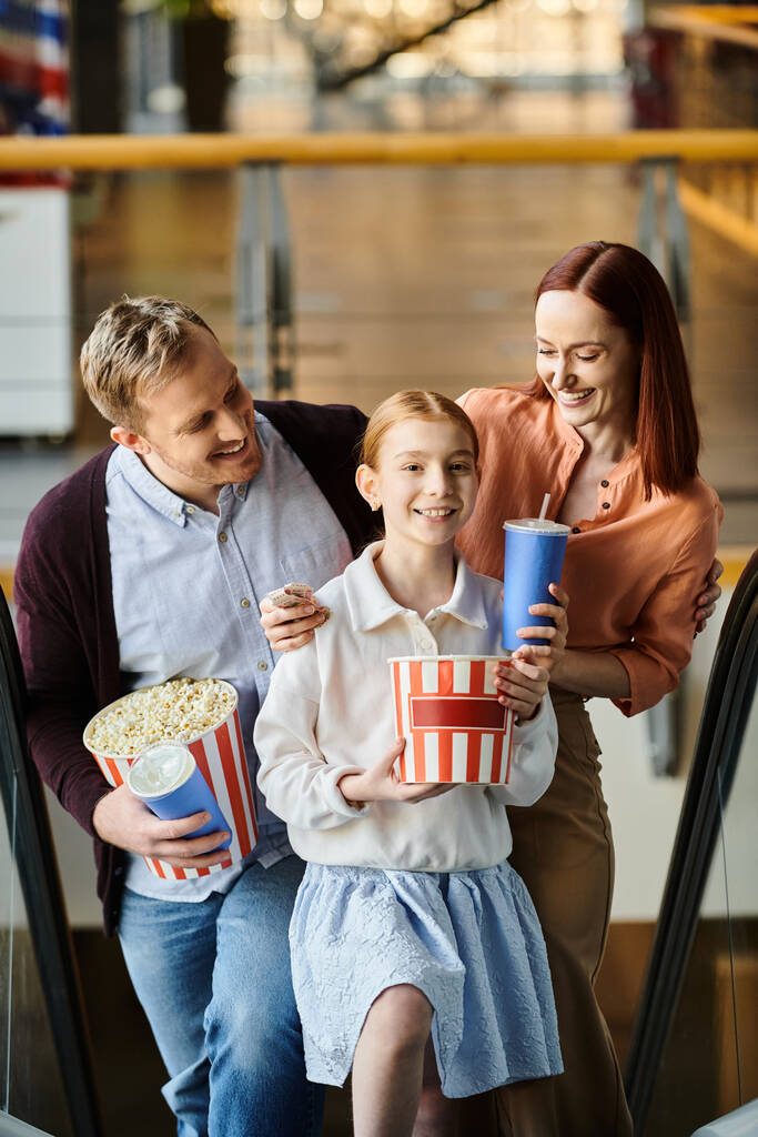 Une famille heureuse, serrant du pop-corn et des boissons, s'assoit sur un escalier roulant, profitant d'une sortie cinéma ensemble. - Photo, image