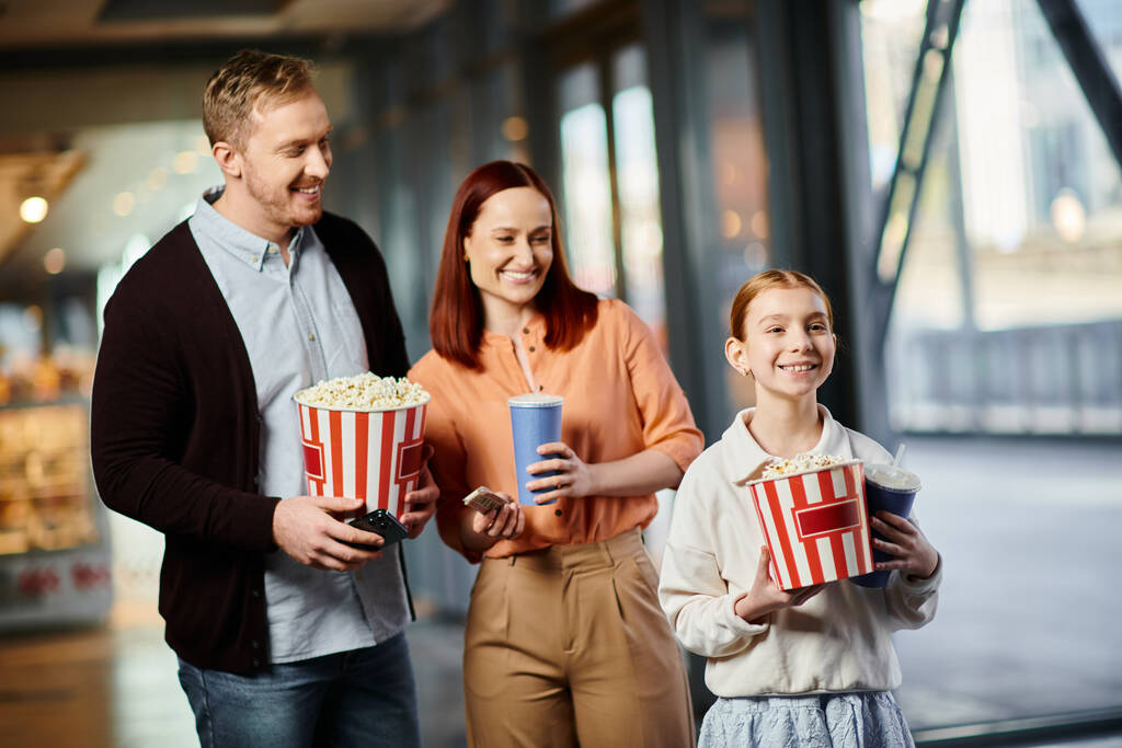 Ένας άντρας, μια γυναίκα και ένα παιδί που κρατάνε χαρούμενα κουτιά ποπ κορν, απολαμβάνοντας μια οικογενειακή έξοδο στο σινεμά.. - Φωτογραφία, εικόνα