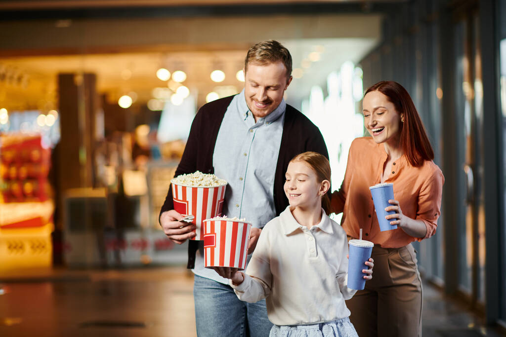 Mężczyzna, kobieta i dziecko szczęśliwie trzymający popcorn w kinie, cieszący się dobrym czasem spędzonym razem. - Zdjęcie, obraz