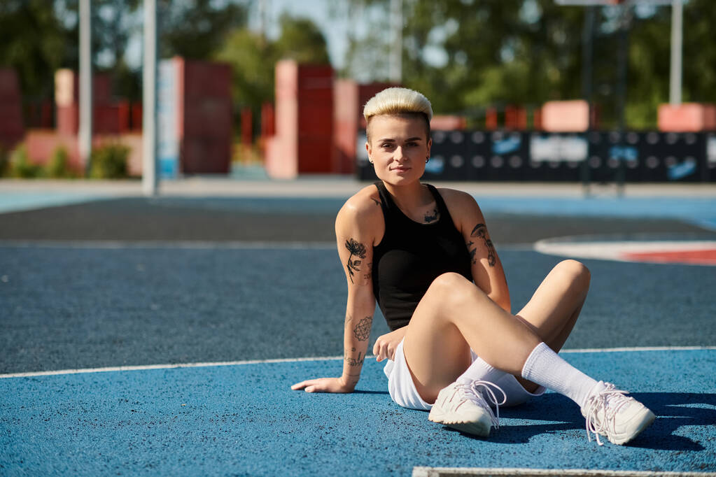 Een jonge vrouw met kort haar en tatoeages zit sierlijk op een basketbalveld, haar benen elegant gekruist. - Foto, afbeelding