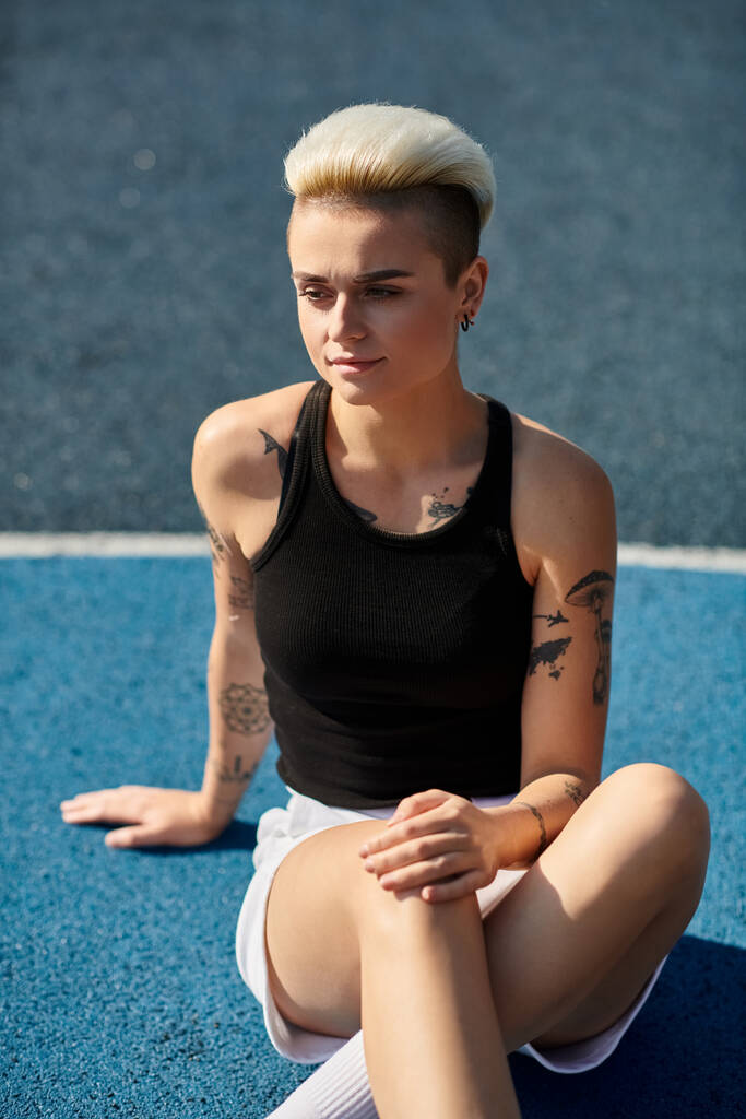 Una joven con el pelo corto y tatuajes sentada con las piernas cruzadas en el suelo en un entorno al aire libre sereno. - Foto, Imagen