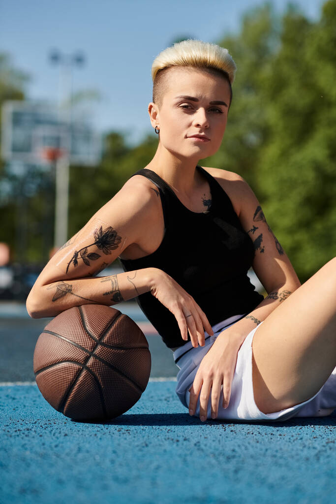 Une jeune femme tatouée assise sur le sol, tenant un ballon de basket, perdue dans ses pensées. - Photo, image