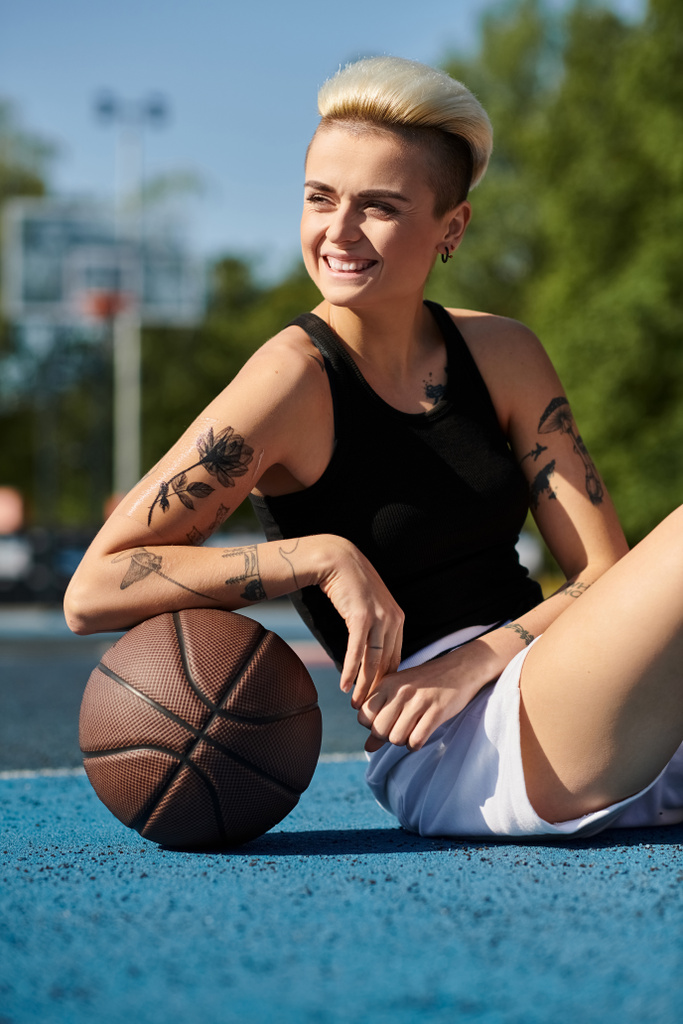 Een jonge vrouw met kort haar en tatoeages zit op de grond, met een basketbal in een stedelijke buitenomgeving. - Foto, afbeelding
