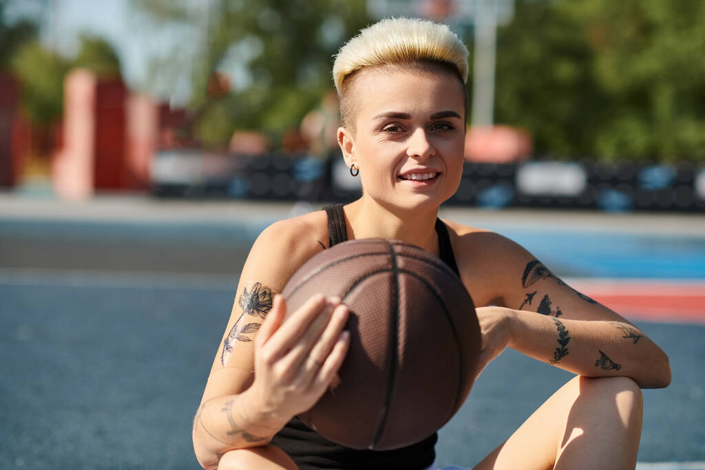 Kısa saçlı ve dövmeli genç bir kadın yere oturmuş, kararlı bir bakışla basketbol topunu sallıyor.. - Fotoğraf, Görsel