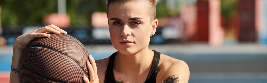 Une jeune femme aux cheveux courts et tatoués tient avec confiance un ballon de basket dans sa main droite, prête à mettre en valeur ses compétences athlétiques à l'extérieur. - Photo, image