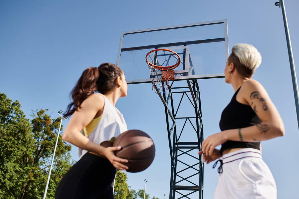 Deux jeunes femmes athlétiques se tiennent ensemble, tenant un ballon de basket, prêtes à jouer à un jeu en plein air par une journée ensoleillée. - Photo, image