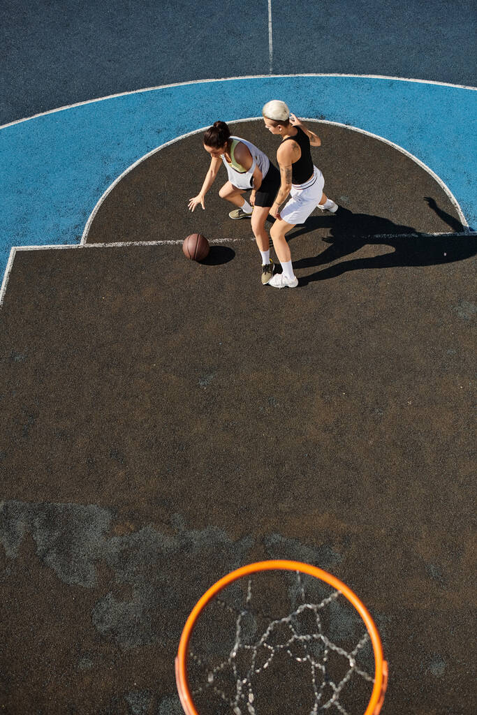 Δύο νεαρές γυναίκες με αθλητικά παίζουν παθιασμένα μπάσκετ σε ένα ηλιόλουστο υπαίθριο γήπεδο.. - Φωτογραφία, εικόνα