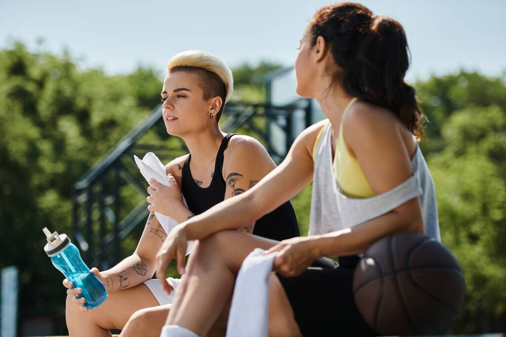 Δύο νεαρές γυναίκες κάθονται μαζί στο γήπεδο του μπάσκετ, κάνουν ένα διάλειμμα από το παιχνίδι, επιδεικνύοντας συντροφικότητα και φιλία. - Φωτογραφία, εικόνα