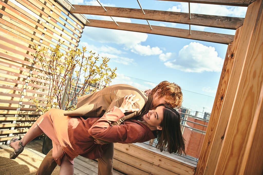 Un homme et une femme s'embrassent paisiblement sur une terrasse en bois surplombant la nature, leurs silhouettes se mélangeant à la lumière du coucher de soleil chaud - Photo, image