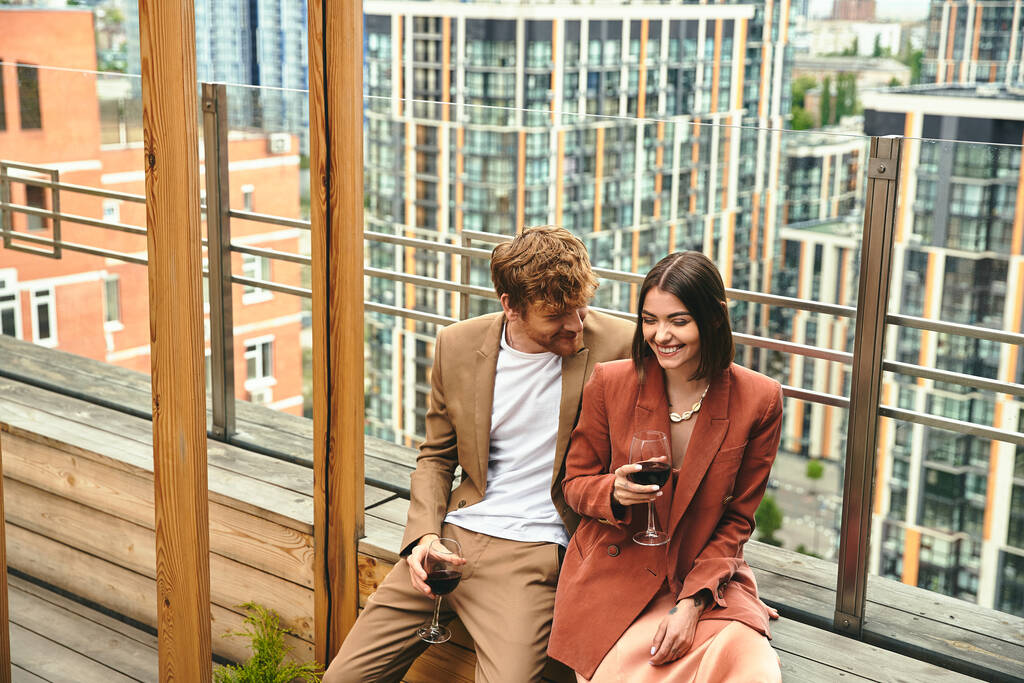 Pár sdílí okamžik smíchu a červeného vína, zarámovaný výhledem na město na dřevěném balkóně - Fotografie, Obrázek
