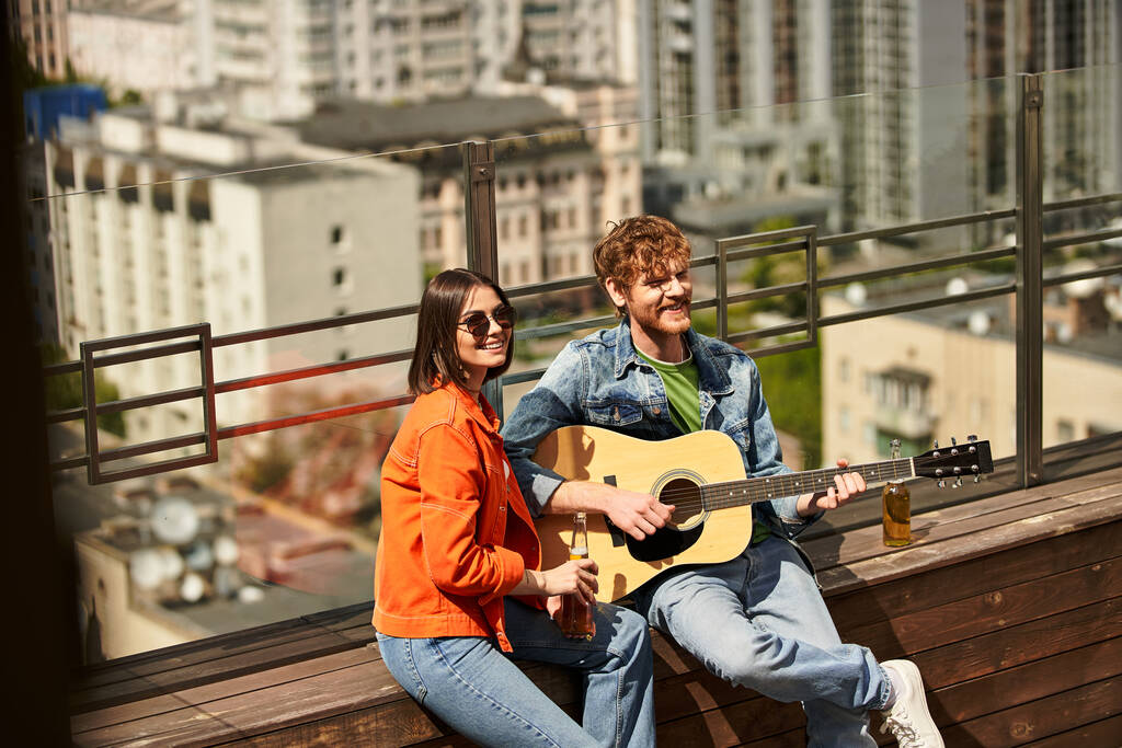 Um homem e uma mulher sentam-se em um banco, ela segura uma guitarra enquanto ele ouve atentamente. Eles compartilham melodias sob o céu aberto - Foto, Imagem