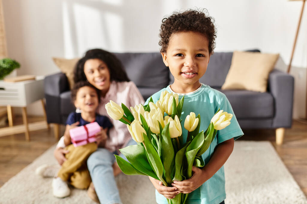 зосередитися на веселому афроамериканському хлопчику, який тримає тюльпани зі своєю розмитою мамою і братом на тлі - Фото, зображення