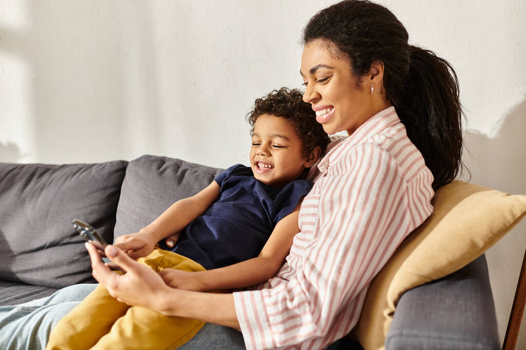 hilpeä kaunis afrikkalainen amerikkalainen äiti ja hänen pieni poikansa katselee puhelinta istuessaan sohvalla - Valokuva, kuva