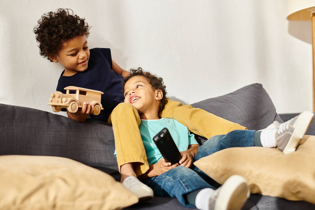 веселые африканские американские мальчики сидят на диване, играя с игрушечной машиной и смотрят друг на друга - Фото, изображение