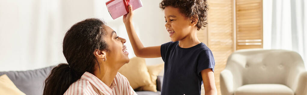 χαριτωμένο αφροαμερικανό αγόρι δίνει δώρο και καρτ ποστάλ στην χαρούμενη μητέρα του, σύγχρονη ανατροφή των παιδιών, πανό - Φωτογραφία, εικόνα