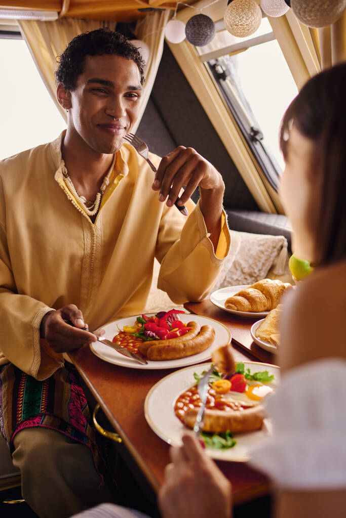 Una pareja interracial disfrutando de un almuerzo romántico juntos en una acogedora caravana con platos de deliciosa comida en la mesa. - Foto, imagen