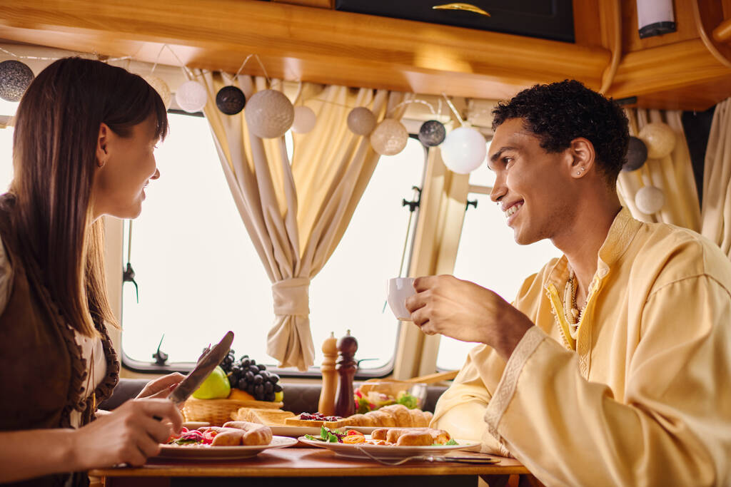Een interraciale man en vrouw zitten aan een tafel, genieten van een heerlijke maaltijd samen in een camper op hun romantische uitje. - Foto, afbeelding