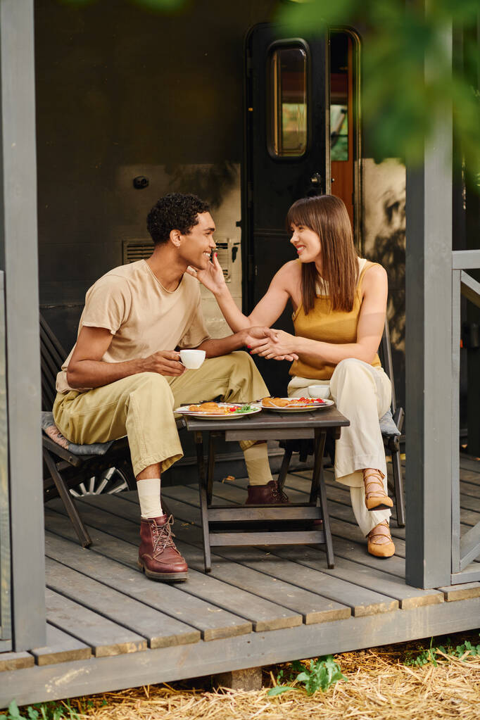 Ένα διαφυλετικό ζευγάρι απολαμβάνει ένα ευχάριστο γεύμα μαζί σε ένα τραπέζι έξω, που περιβάλλεται από τη φύση και τη ζεστή αγκαλιά της αγάπης. - Φωτογραφία, εικόνα
