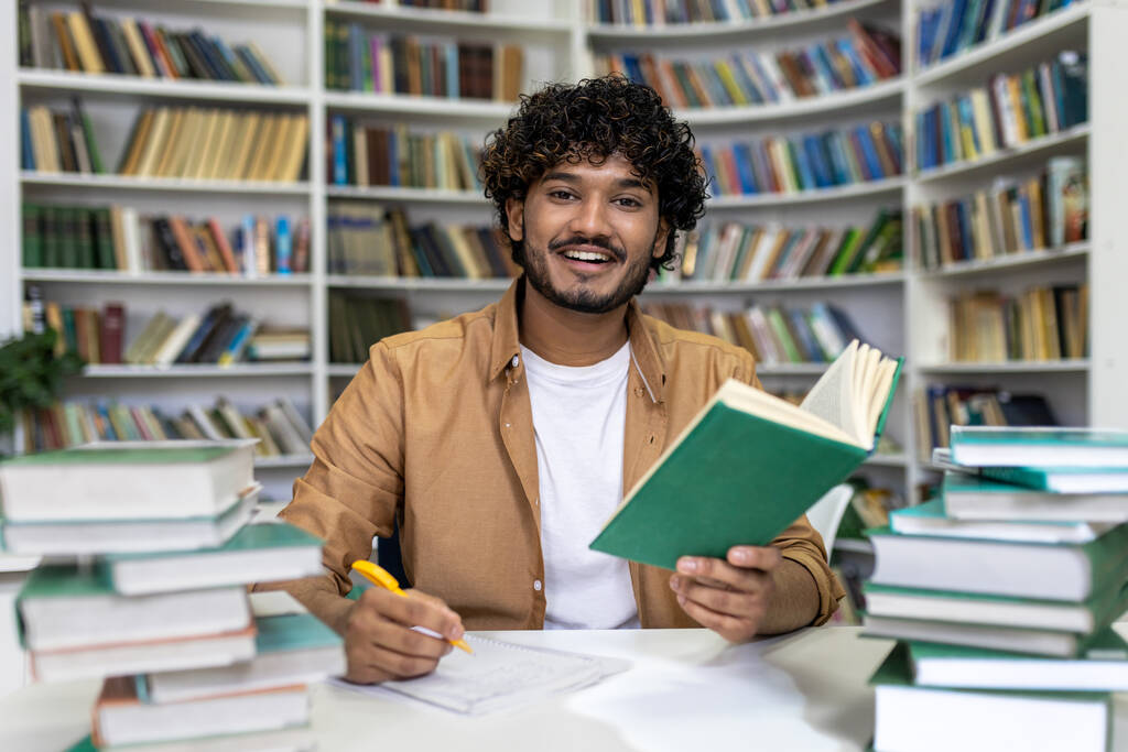 Szczęśliwy młody student przygotowujący się do egzaminów w bibliotece uniwersyteckiej. Otoczony przez duże stosy książek, reprezentuje poświęcenie dla sukcesu akademickiego i dążenie do wiedzy w - Zdjęcie, obraz