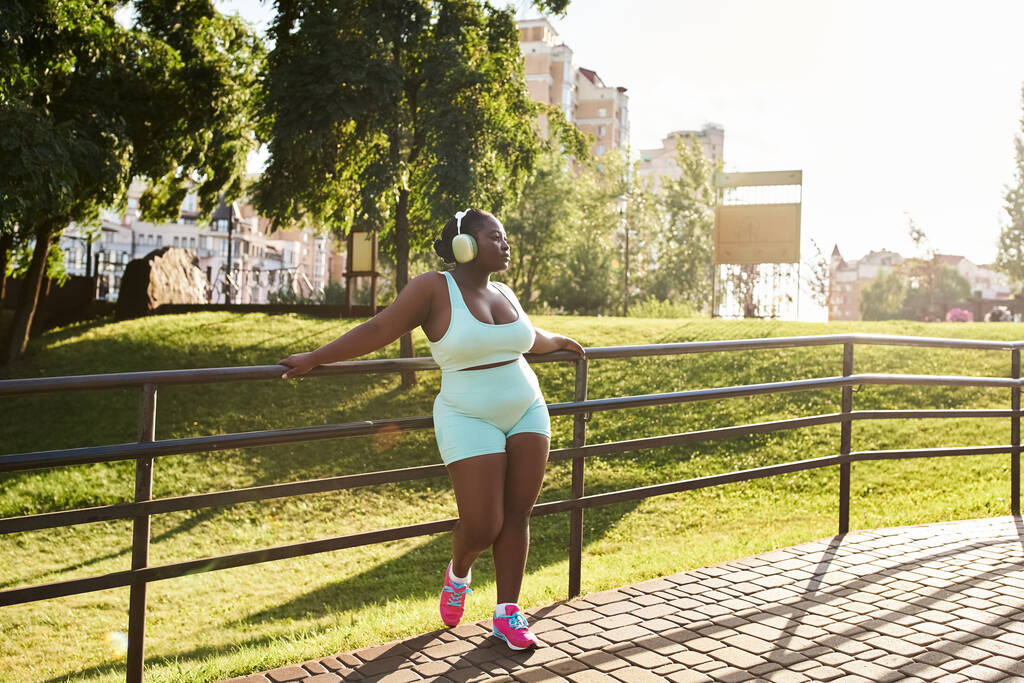 Una mujer afroamericana con curvas en audífonos se apoya en una valla, con un top blanco y pantalones cortos azules, perdidos en la música. - Foto, imagen