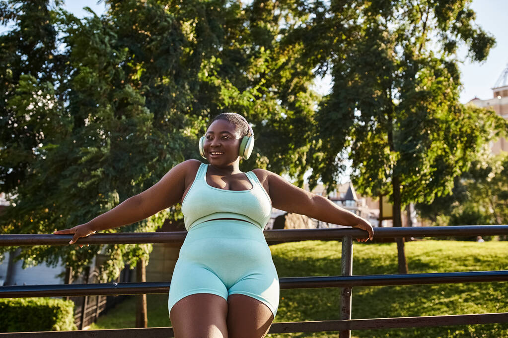An African American woman in blue shorts and headphones leans on a rail, deeply engrossed in her music. - Photo, Image