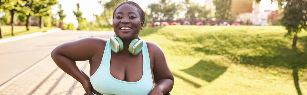 Uma mulher afro-americana elegante com uma figura curvilínea, vestindo um top azul e fones de ouvido, desfrutando de música ao ar livre. - Foto, Imagem