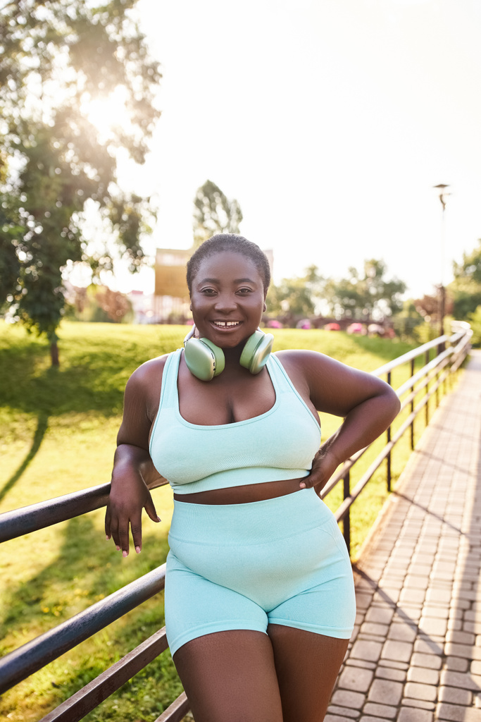 Afrikkalainen amerikkalainen nainen kuulokkeet, yllään sininen toppi ja shortsit, luottavaisesti poseeraa kuvan ulkona. - Valokuva, kuva