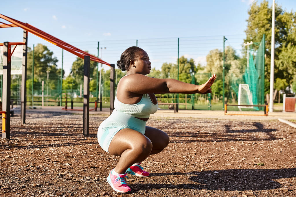 Игривая афроамериканка в спортивной одежде, приседающая в парке с детской площадкой на заднем плане. - Фото, изображение