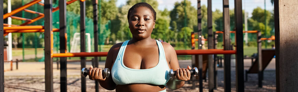 Μια Αφρο-Αμερικανίδα με καμπυλωτό σώμα γυμνάζεται με ένα μπλε αθλητικό σουτιέν, κρατώντας αλτήρες μπροστά σε μια παιδική χαρά. - Φωτογραφία, εικόνα