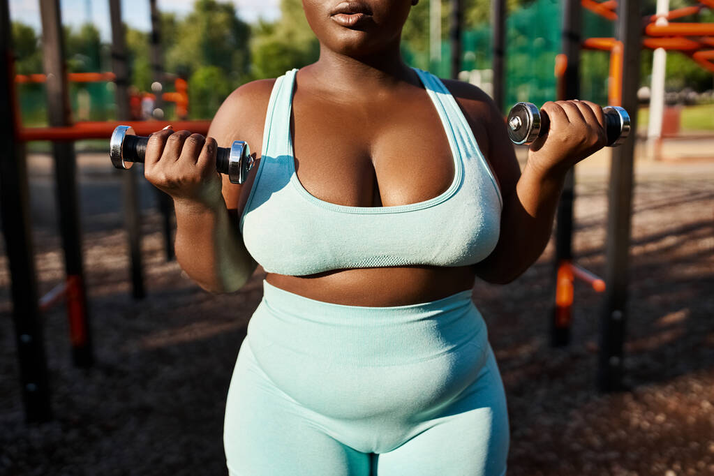 Egy testpozitív afro-amerikai nő kék sportruhában magabiztosan felemel két súlyzót a szabadban.. - Fotó, kép