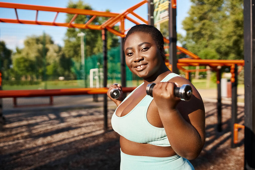 Een Afro-Amerikaanse vrouw in sportkleding oefent zelfverzekerd met een halter in een weelderig park, omarmt lichaamspositiviteit. - Foto, afbeelding