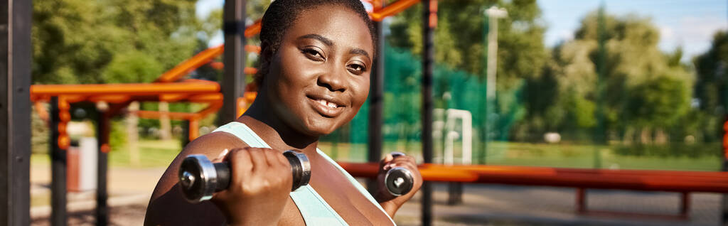 緑豊かな公園の設定でダンベルとスポーツウェアのアフリカ系アメリカ人女性. - 写真・画像