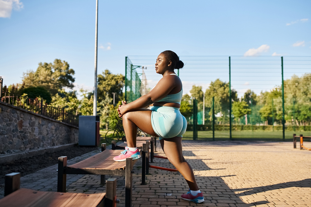 Une femme afro-américaine en tenue de sport contemple alors qu'elle s'entraîne gracieusement sur un banc en bois à l'extérieur. - Photo, image