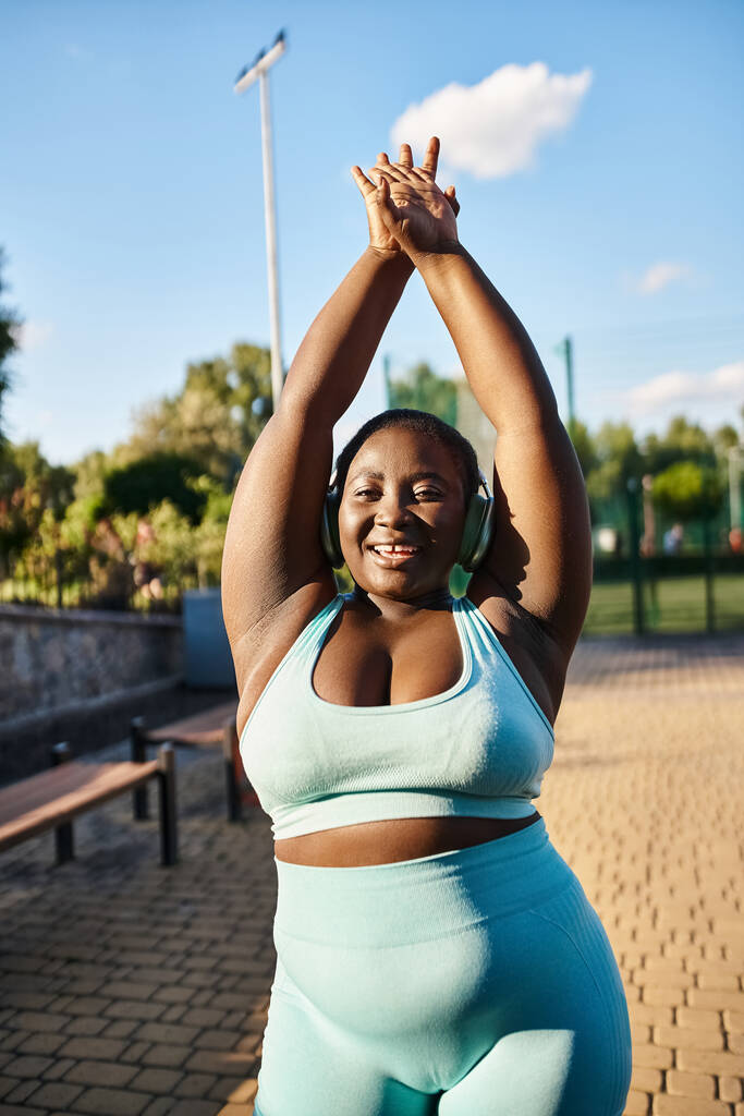 Une femme afro-américaine avec une attitude positive de corps, portant un haut de soutien-gorge de sport bleu, étend ses bras gracieusement à l'extérieur. - Photo, image