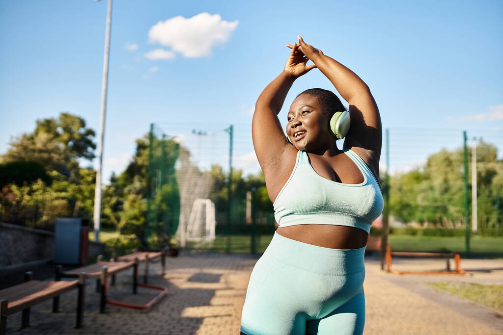 Mulher afro-americana curvilínea no top de sutiã esportivo estende os braços ao ar livre, promovendo a positividade do corpo e fitness. - Foto, Imagem