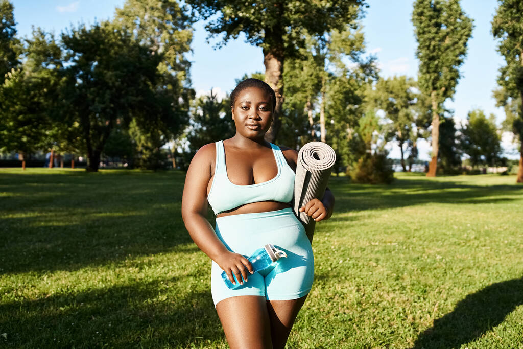Uma mulher afro-americana em sportswear segurando tapete esportivo ao ar livre, incorporando positividade do corpo e força. - Foto, Imagem