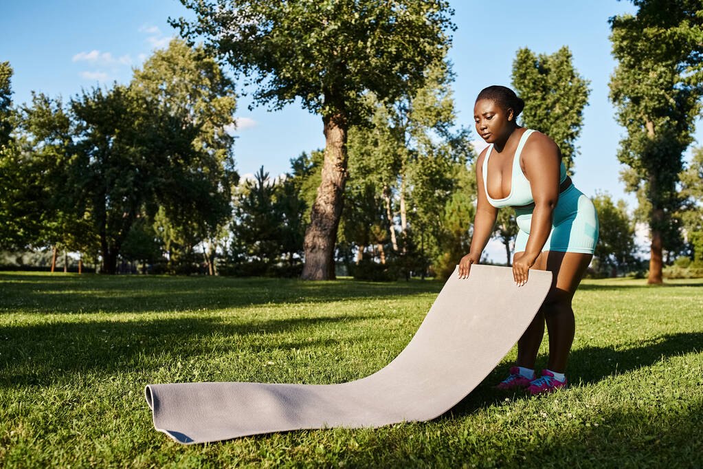 Μια καμπυλωτή Αφρο-Αμερικανίδα γυναίκα με αθλητικά να στέκεται με αυτοπεποίθηση δίπλα σε ένα αθλητικό χαλάκι. - Φωτογραφία, εικόνα