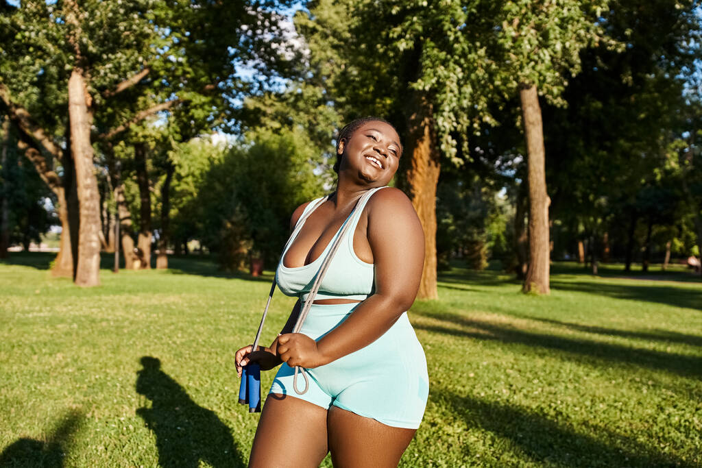Afroamerikkalainen nainen urheiluvaatteissa seisoo kauniisti pellolla korkeiden puiden ympäröimänä säteillen kehon positiivista auraa.. - Valokuva, kuva