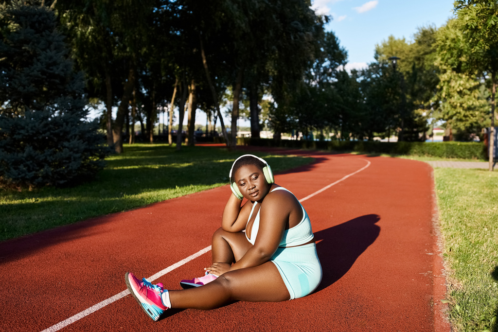 Afrikkalainen amerikkalainen nainen urheiluvaatteissa istuu tenniskentällä, pitäen mailaa valmistellessaan peliä.. - Valokuva, kuva