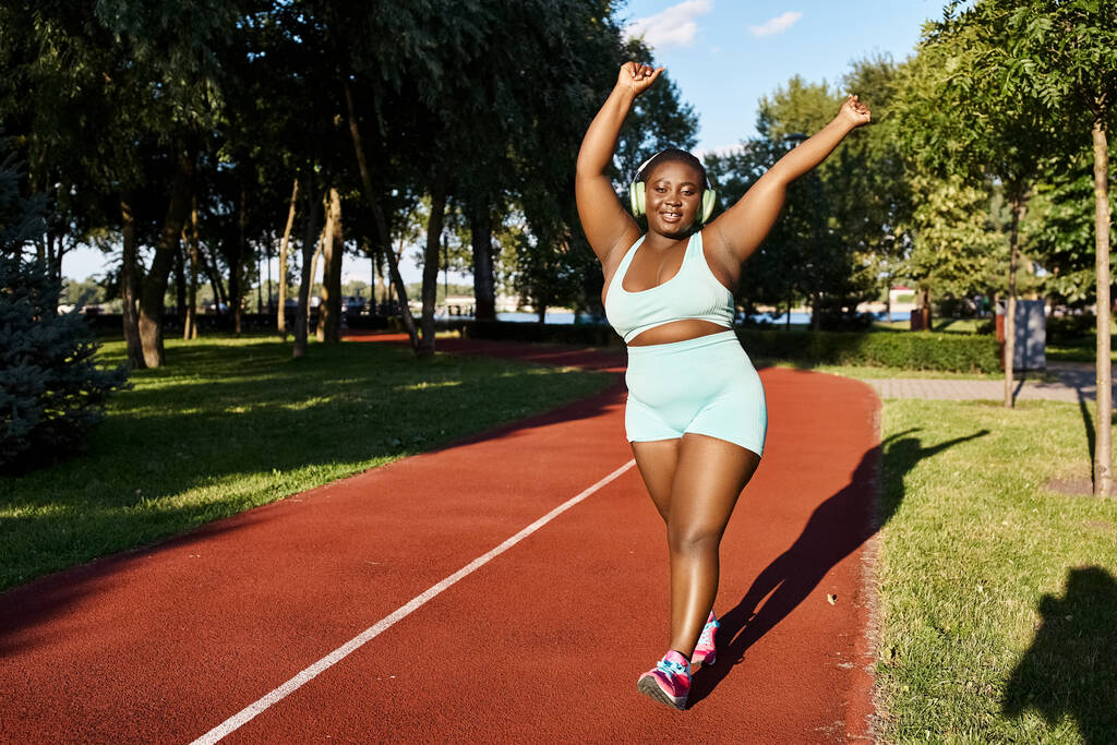 Uma mulher afro-americana em sportswear corre em uma pista com árvores no fundo, mostrando sua forma corpo-positiva e curvilínea. - Foto, Imagem