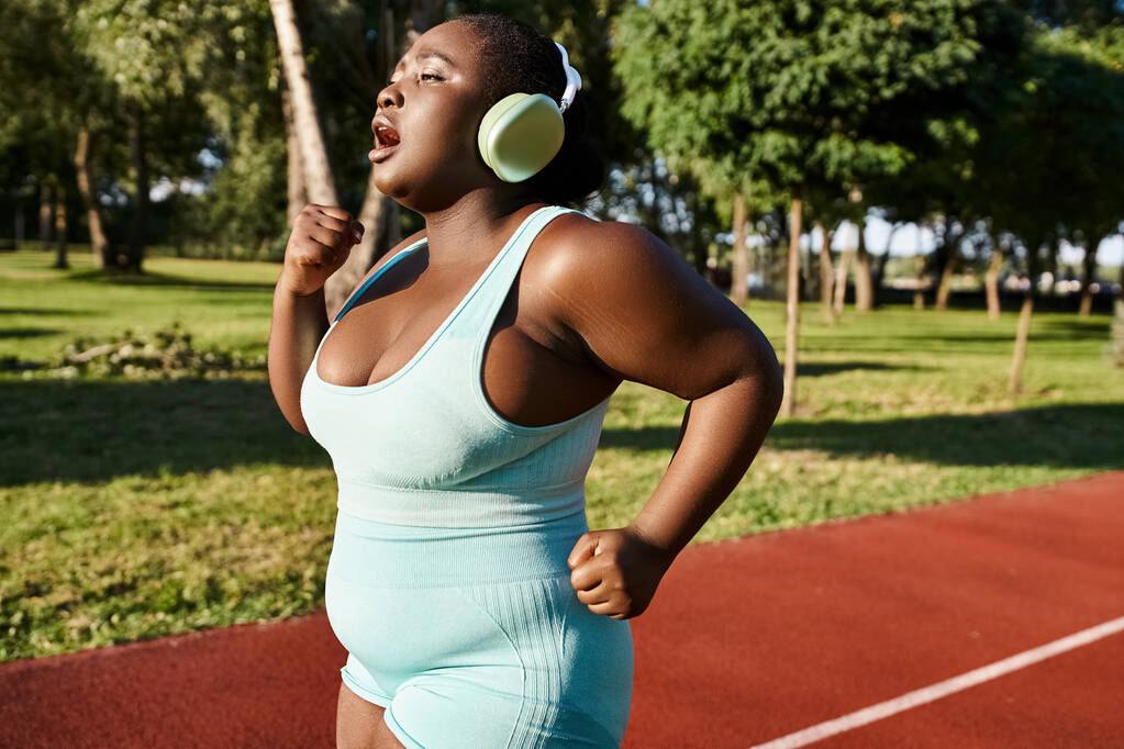 Uma afro-americana positiva para o corpo em roupas esportivas fica em uma quadra de tênis usando fones de ouvido enquanto desfruta de música. - Foto, Imagem
