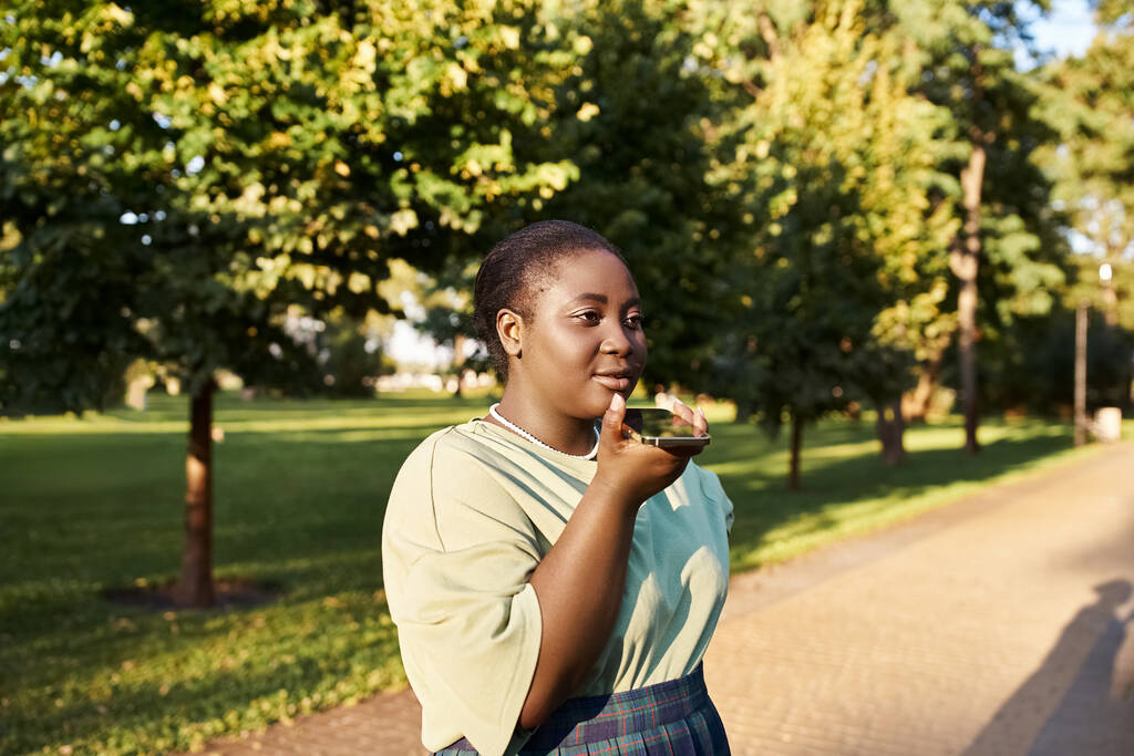 Plus-size donna afroamericana si trova sul ciglio della strada, chiacchierando al telefono in estate caldo, abbracciando positività del corpo. - Foto, immagini