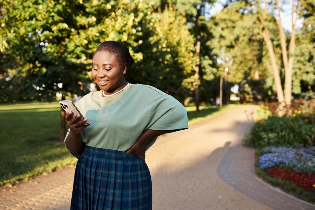 Plus size donna afroamericana in abbigliamento casual in piedi in un parco, guardando il suo telefono cellulare in una giornata di sole. - Foto, immagini