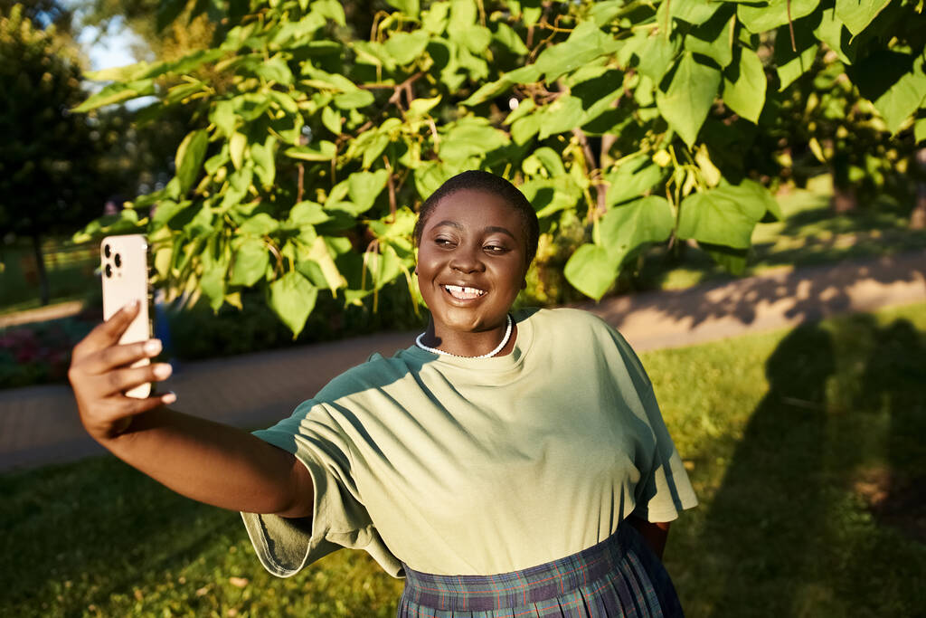 Uma mulher afro-americana plus-size em traje casual alegremente tirar uma selfie com seu telefone celular ao ar livre no verão. - Foto, Imagem