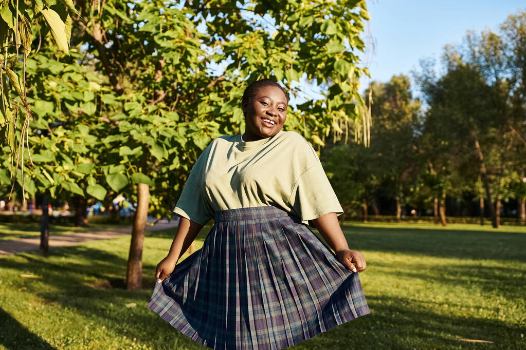 Une femme afro-américaine de taille plus se tient gracieusement dans l'herbe d'été, portant une jupe fluide et exsudant la positivité du corps. - Photo, image