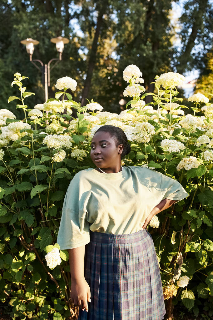 Eine Afroamerikanerin im Plus-Size-Format steht selbstbewusst vor einem Strauch mit weißen Blumen und umarmt die Schönheit der Natur um sich herum.. - Foto, Bild