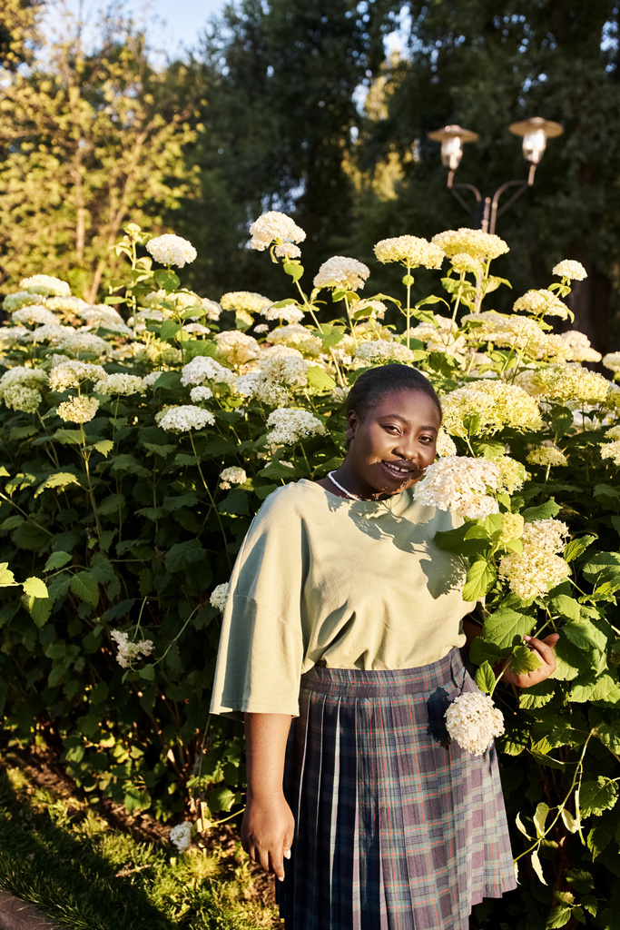 Плюс размер афроамериканка уверенно стоя перед красочным кустом цветов в летнее время. - Фото, изображение
