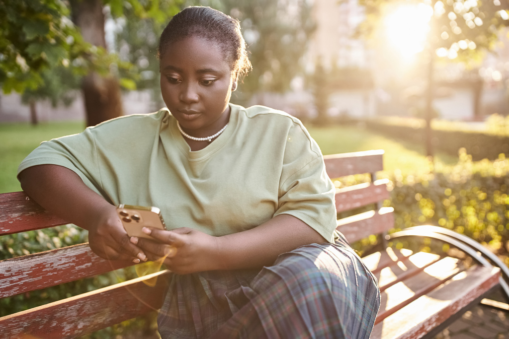 Una donna afro-americana plus-size in abbigliamento casual seduta su una panchina di legno in un parco in una giornata estiva soleggiata, abbracciando la positività del corpo. - Foto, immagini