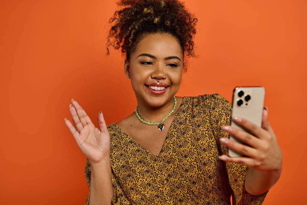 Stilvolle Afroamerikanerin in modischer Kleidung posiert mit einem Handy vor leuchtend orangefarbenem Hintergrund. - Foto, Bild
