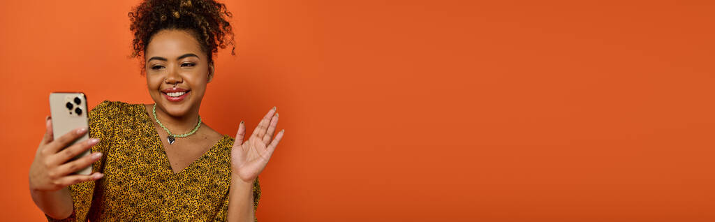 Egy gyönyörű afro-amerikai nő elegáns öltözékben egy mobilt tart egy narancssárga fal előtt.. - Fotó, kép
